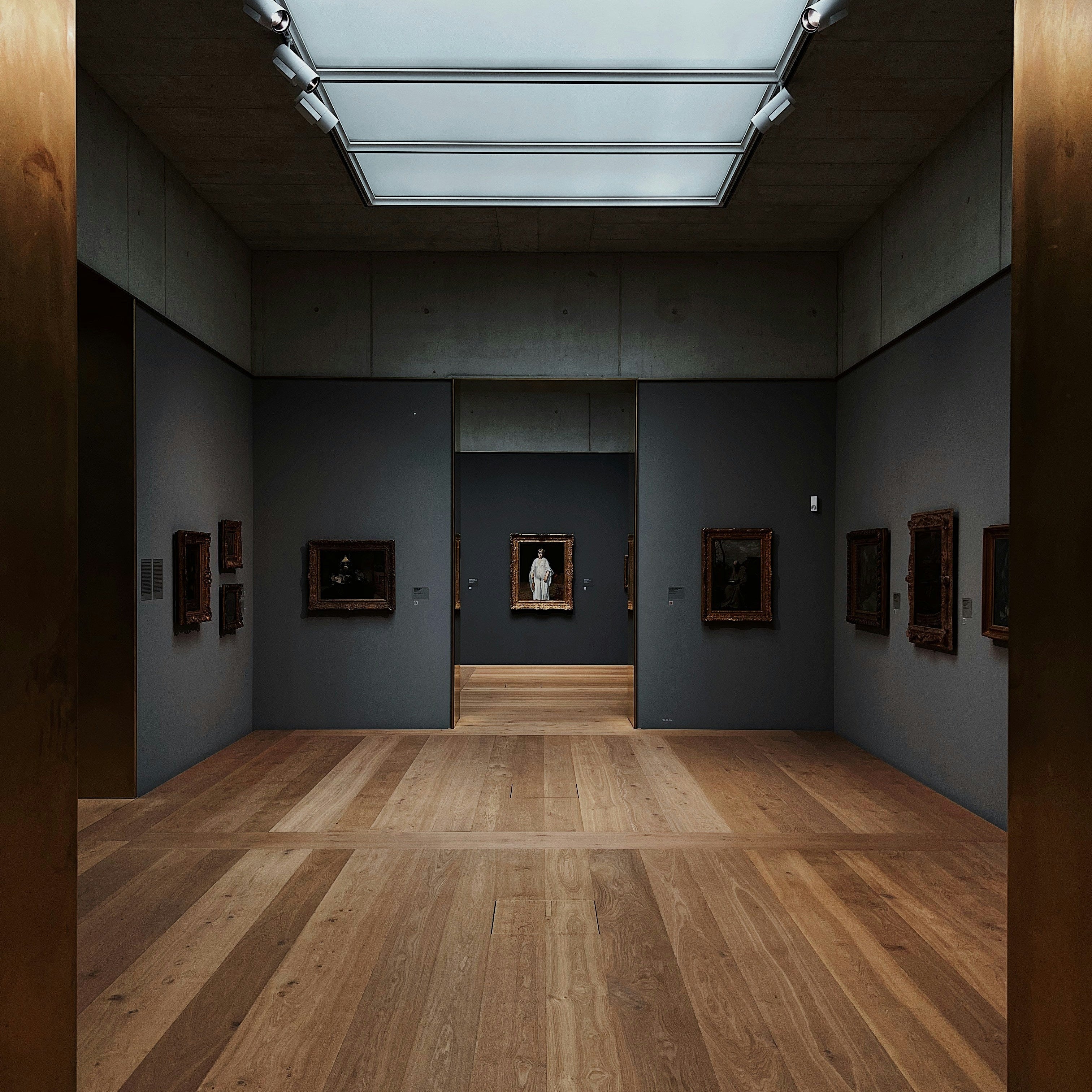 A long gallery interior with a wood plank floor, dark gray walls, framed artwork, and a large skylight overhead.